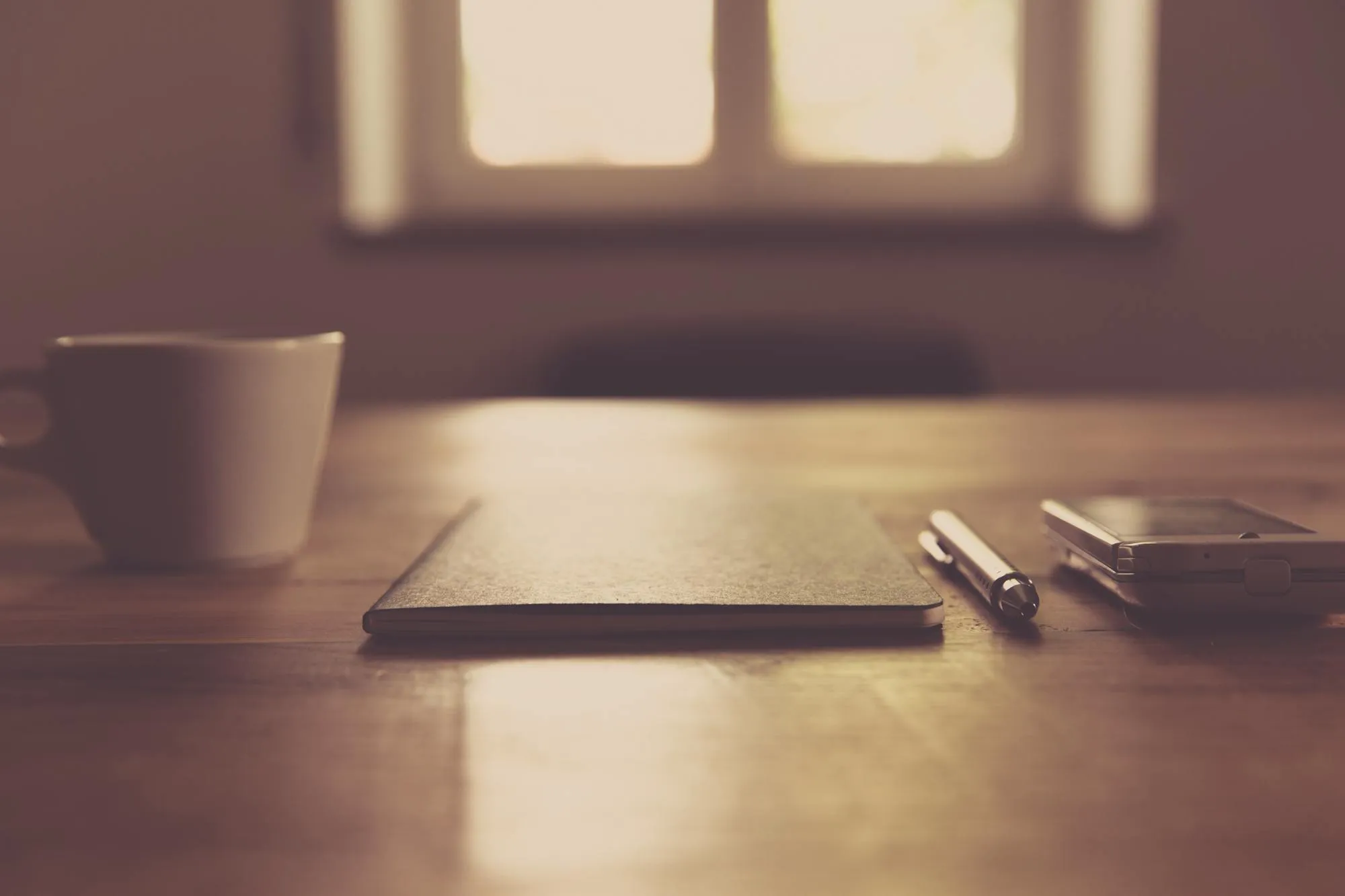 A quiet desk in soft window light: a notebook, a pen, a phone and a cup of coffee